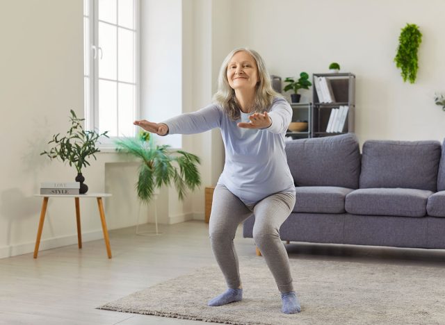 Squat άσκηση της εκπαίδευσης ανώτερων γυναικών στο σπίτι, μπροστινή θέα. Παλιά ευτυχισμένη κυρία Squatting με χέρια που εκτείνονται προς τα εμπρός και χαμόγελο, ενεργό αθλητική προπόνηση σε χαλί στο σαλόνι. Rehab, έννοια ευεξίας