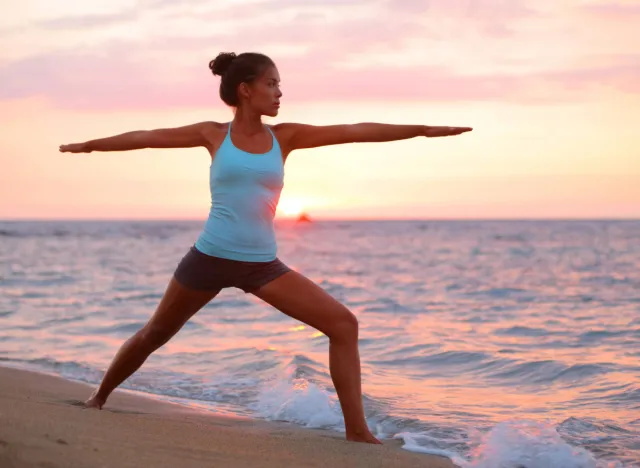 Η Fit Woman που κάνει έναν πολεμιστή θέτει ως μέρος μιας πρωινής προπόνησης γιόγκα στην παραλία κατά την ανατολή του ηλίου
