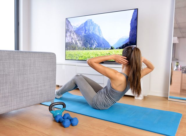 Home Workout - Γυναίκα που ασκεί μπροστά σε μια επίπεδη οθόνη βλέποντας ένα πρόγραμμα γυμναστικής ή ασκώντας κατά τη διάρκεια μιας τηλεοπτικής εκπομπής που βρίσκεται σε ένα χαλάκι γιόγκα μπροστά από τον καναπέ στο σαλόνι ενός σπιτιού ή ενός διαμερίσματος.